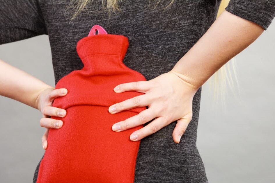A person wearing a grey long-sleeved shirt is putting a hot water bottle on the lower back using both of their hands
