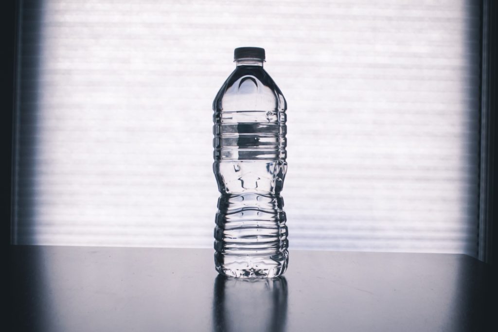 A bottle of distilled water is on the middle of a reflective table and in front of a white textured background