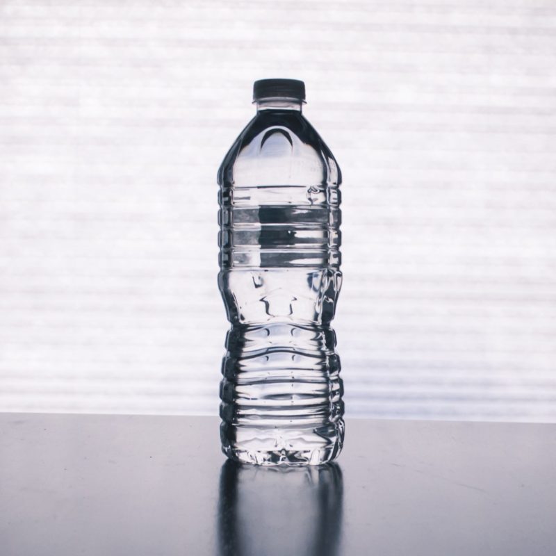 A bottle of distilled water is on the middle of a reflective table and in front of a white textured background