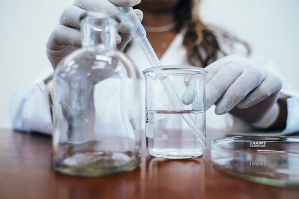 A scientist wearing a white coat and white gloves is pipetting some distilled water while other chemistry glassware is on the wooden table 