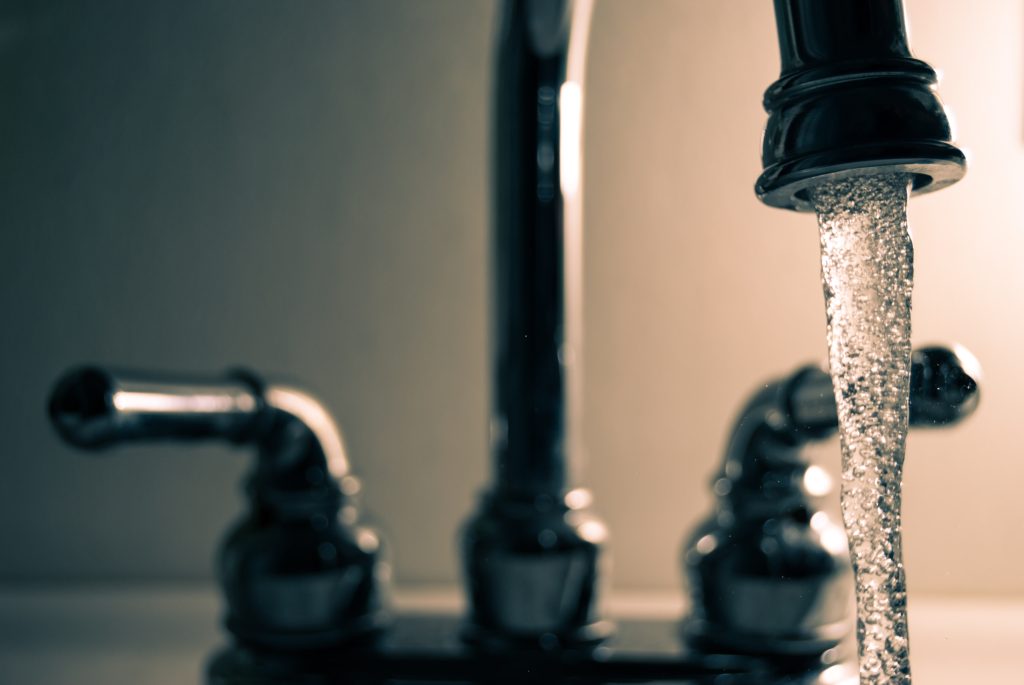 A faucet has the water running while inside a dimly lit room. the water is in focus