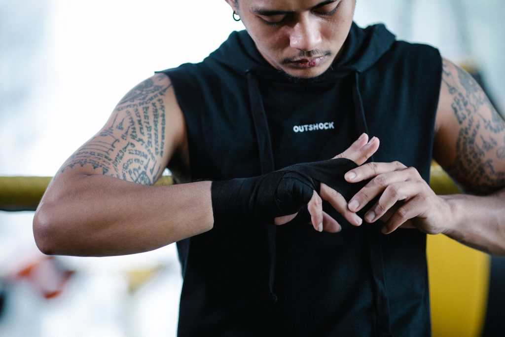 A person with arm tattoos wearing a short-sleeved black hoodie in a gym is pulling at the wrap on their hands, possibly considering preventative athletic taping in the future