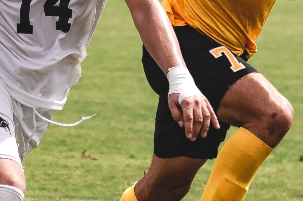 Two pro athletes on the field are in frame; one is wearing a yellow jersey shirt and black jersey shorts while the other one is wearing a full white set which includes sports medicine taping on one of their wrists for preventative measures