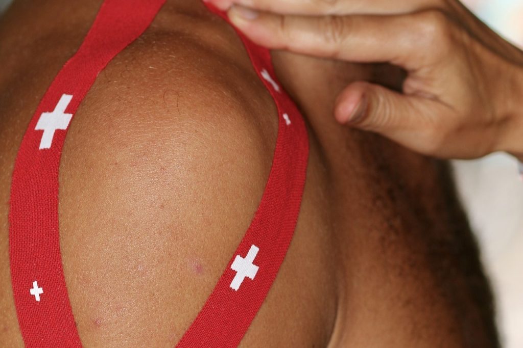 A person is getting sports medicine taping on their shoulder by a physiotherapist using an athletic tape with a red background and a white cross design