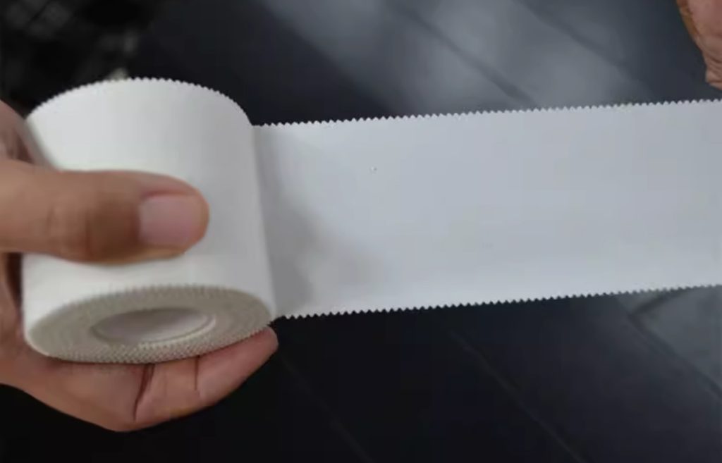 A person is unrolling white sports medicine tape and showing its strength and edges quite clearly 