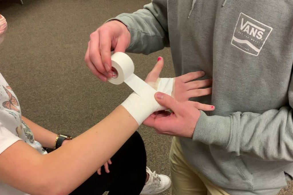 An athlete wearing a white shirt is getting their wrist taped up with preventative athletic taping by a physiotherapist wearing a grey hoodie