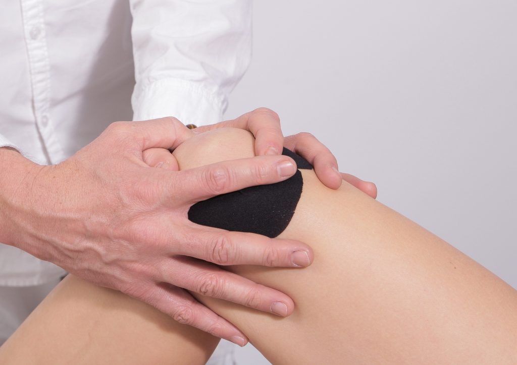 A physiotherapist is putting on some black sports medicine tape as a preventative measure for a patient's knee while wearing a white coat in front of a light grey background