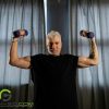 A muscular older gentleman holding small barbells in his hands while he flexes.
