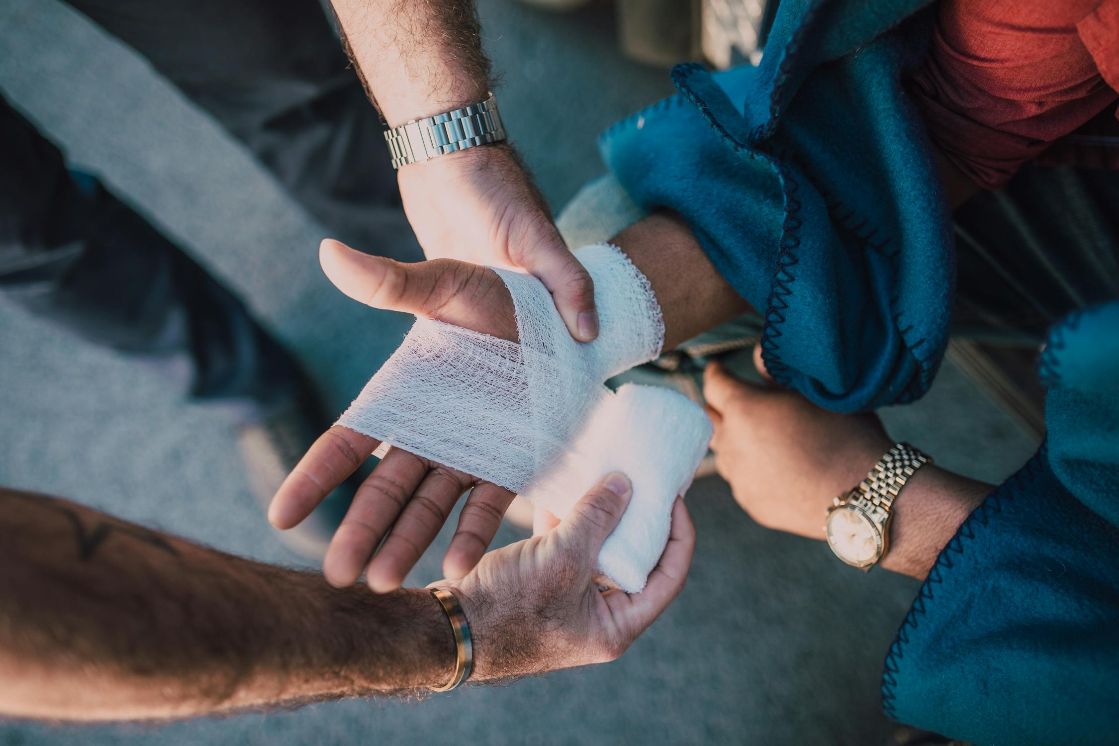 A close-up photo of two pairs of hands. The pair of hands on the left is providing first aid by bandaging an outstretched right hand on the right side of the image.