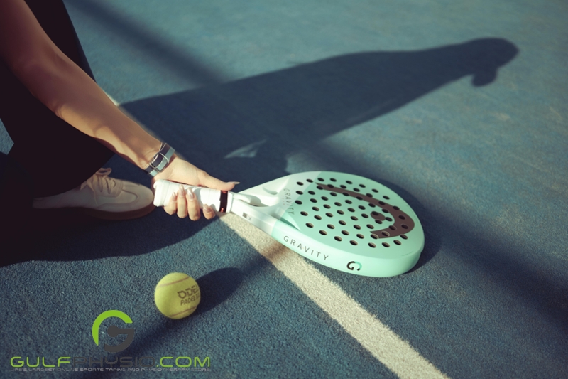 A padel racket with a hand holding it out of frame. There is a game ball on the court.