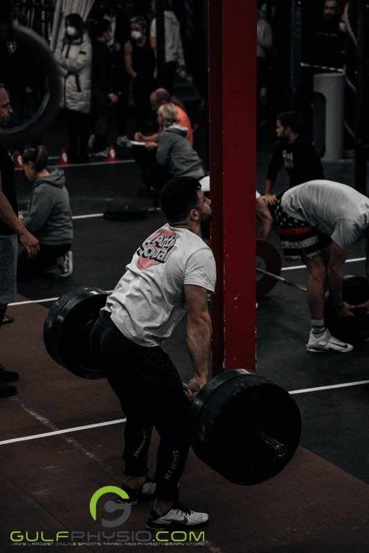 This photo captures multiple people lifting barbells with heavy weights. There are noticeable groups of people in the background, some of them wearing face masks.