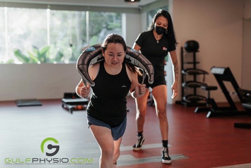 A woman appears to be engaging in the exercise weighted walking lunges inside a fitness center. She is closely monitored by her trainer.